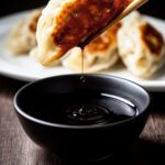 Pan-fried potsticker held by chopsticks — soy sauce dripping into bowl below, steaming hot from the pan